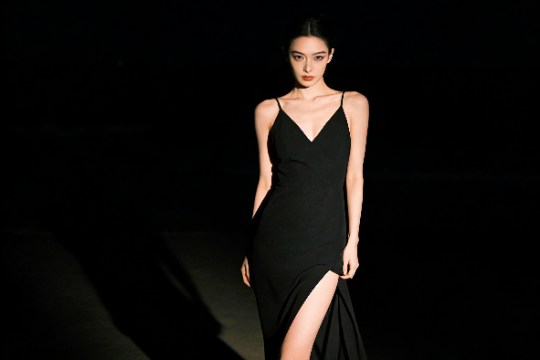 Woman in elegant black dress with thigh-high slit posing against dark background.
