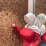 Woman in hijab, red top, and jeans poses against a textured wall, reflected in a mirror.