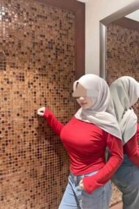 Woman in hijab, red top, and jeans poses against a textured wall, reflected in a mirror.