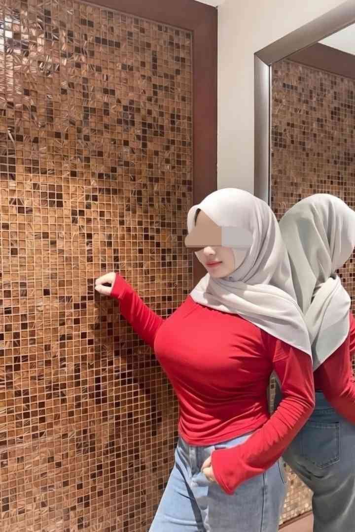 Woman in hijab, red top, and jeans poses against a textured wall, reflected in a mirror.