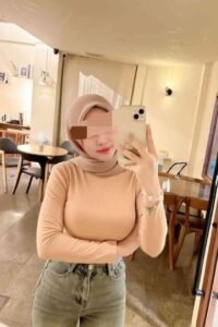 Woman in hijab takes selfie in cafe, wearing tan top and jeans.