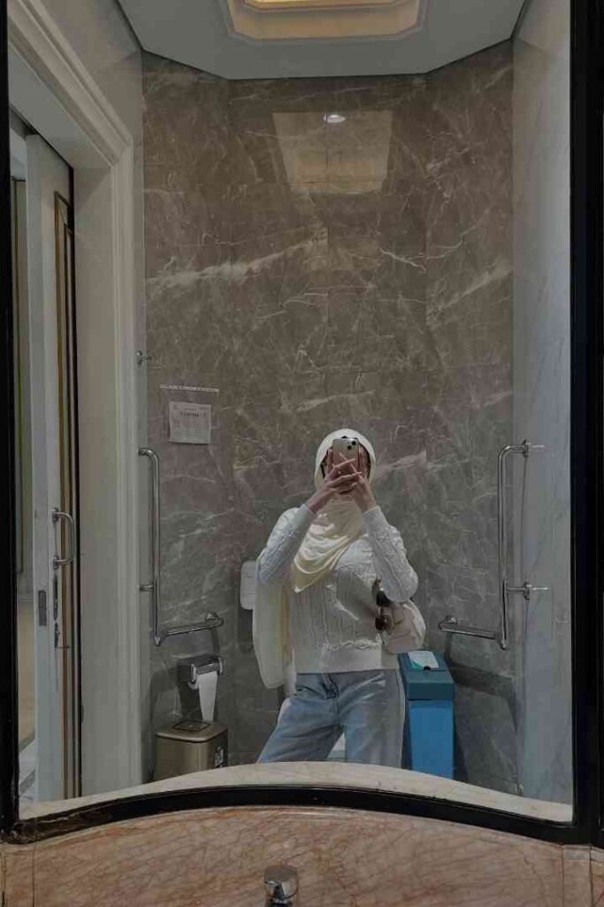 Woman in hijab and white sweater taking mirror selfie in modern bathroom