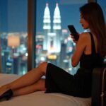 Woman in hotel room with city view using smartphone.